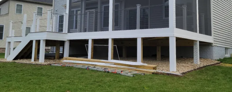A white deck with stairs leads to a screened porch; wooden beams support it, set on a gravel bed in front of a house.