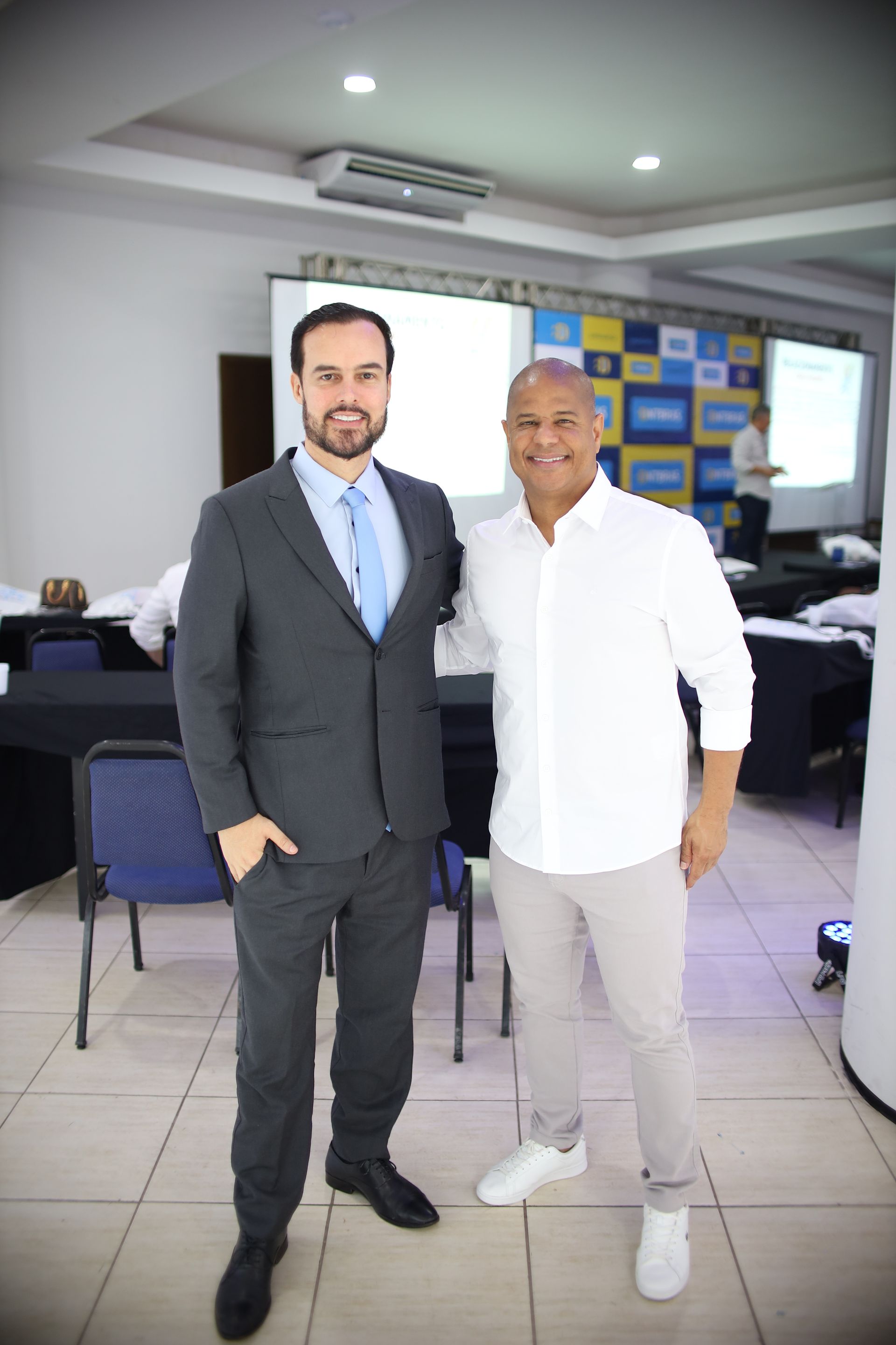 Dois homens posam juntos em uma sala de conferências. Um de terno, o outro de camisa branca e calça, sorrindo.