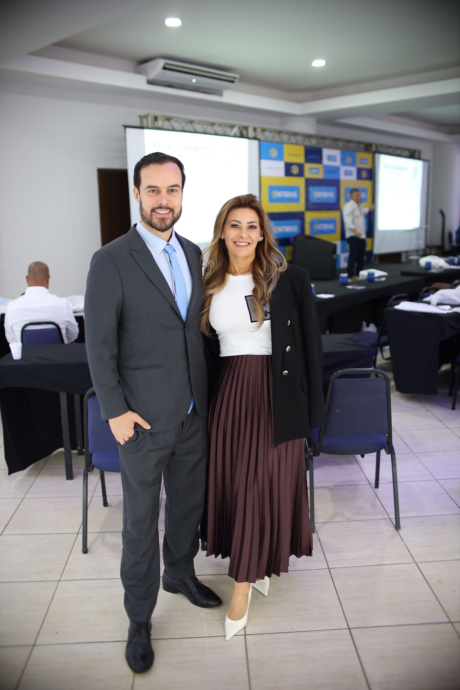 Um homem e uma mulher estão juntos em uma sala de conferências. O homem veste terno, a mulher saia e jaqueta.