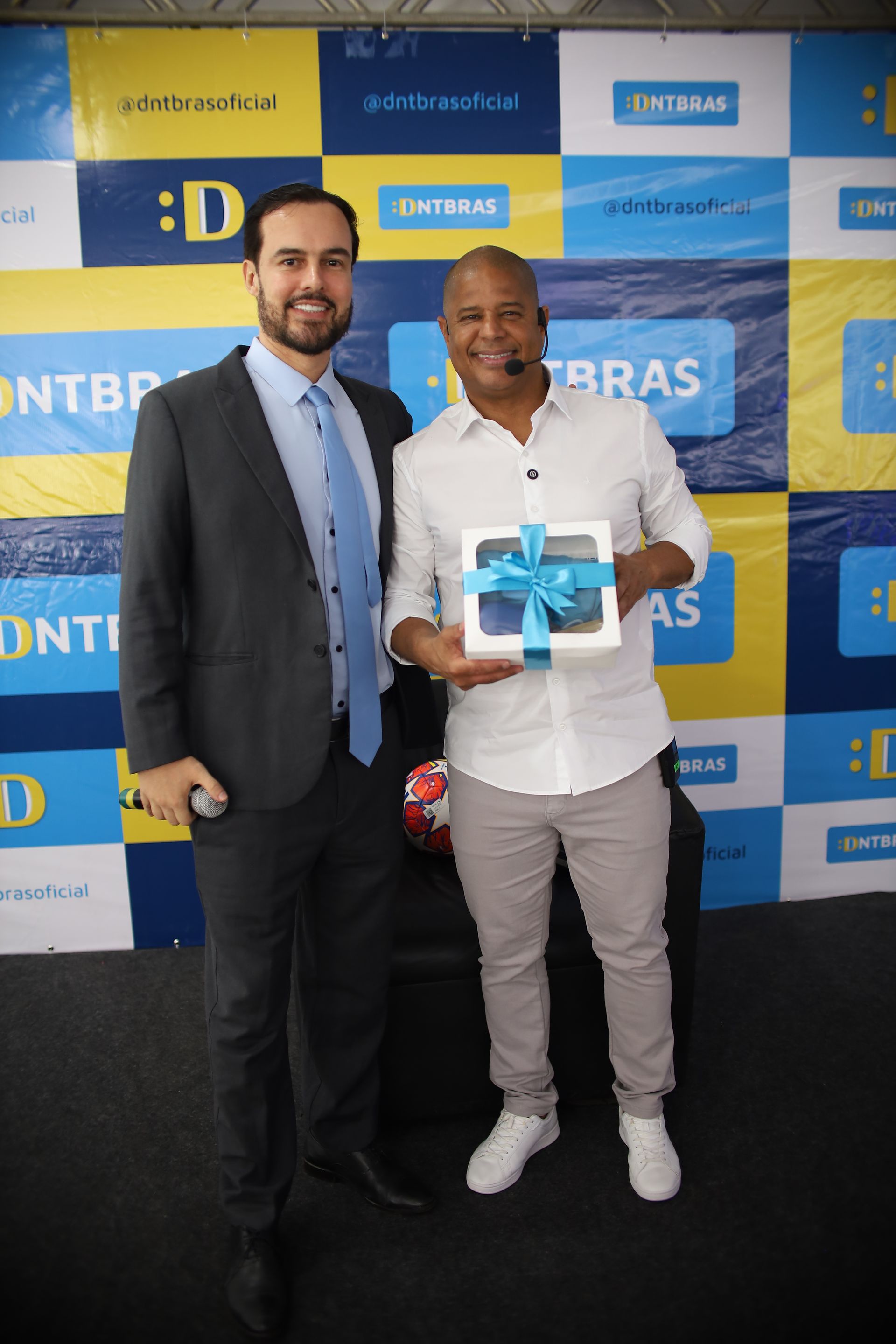 Dois homens posam para uma foto. Um de terno, o outro de camisa branca, segurando uma caixa de presente amarrada com uma fita azul.