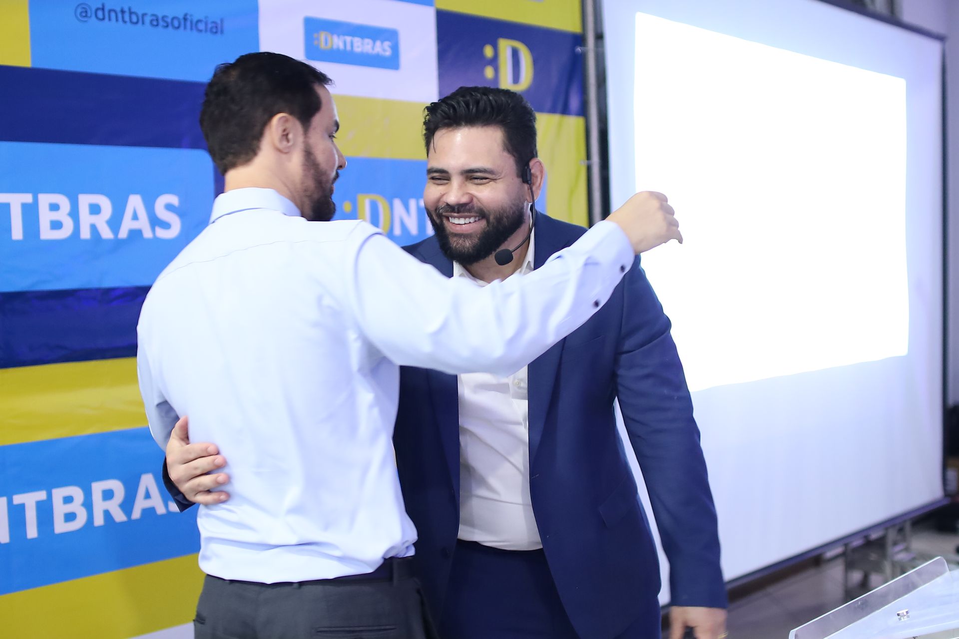 Dois homens se abraçam em um escritório. Um veste terno azul, o outro camisa azul-clara. Tela branca e logotipo ao fundo.