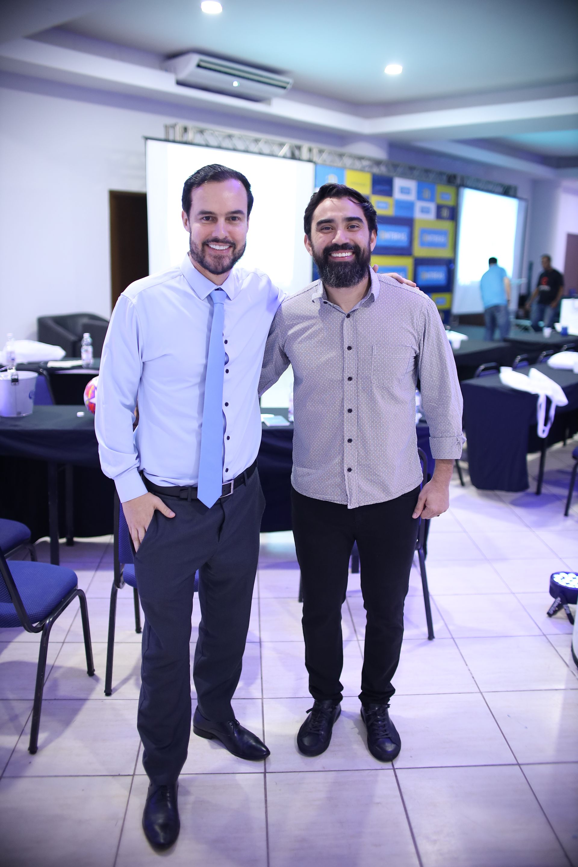 Dois homens posam para uma foto em uma sala de conferências. Um veste camisa azul e gravata, o outro, camisa estampada.