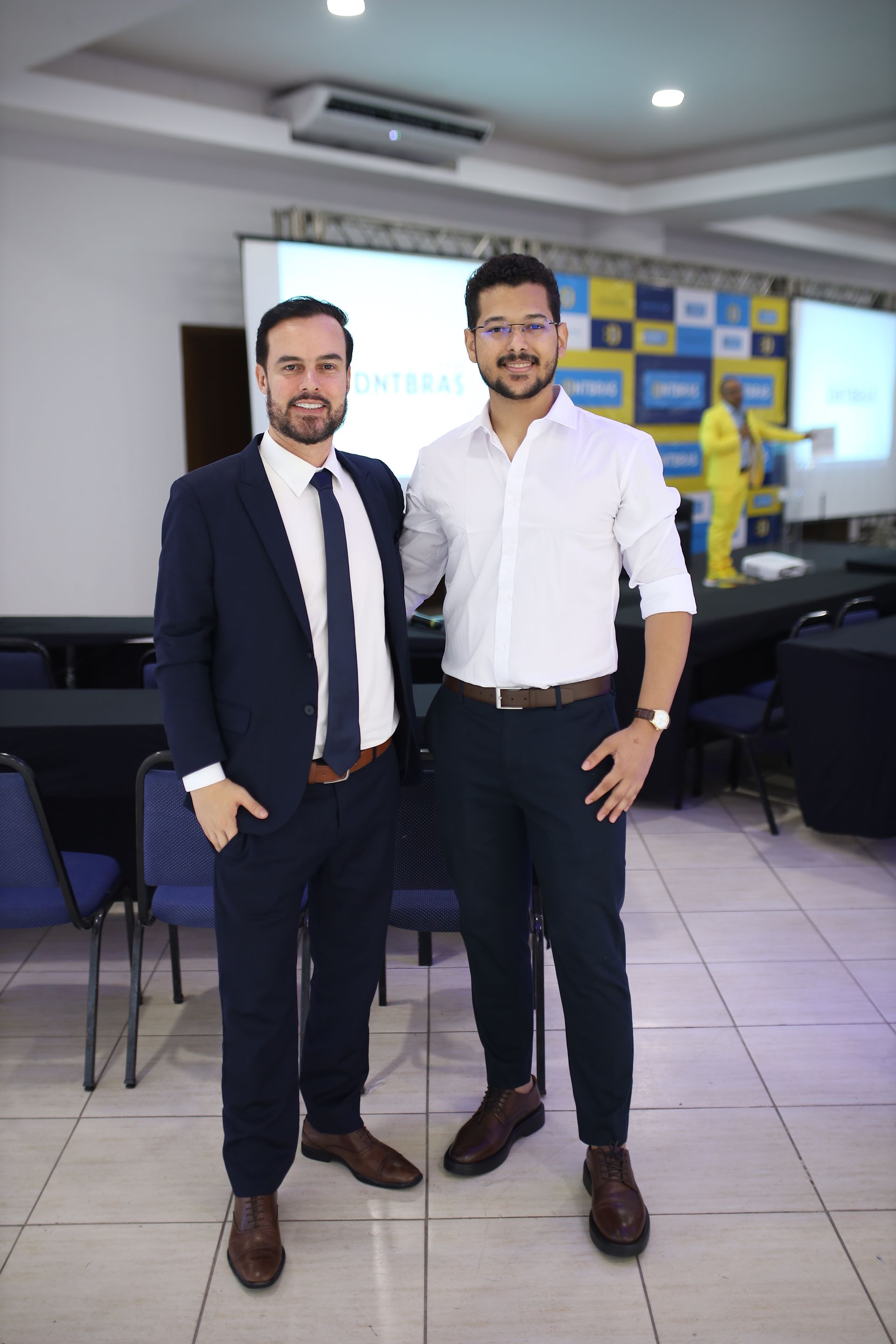 Dois homens em trajes de negócios posam para uma foto em um ambiente de conferência.