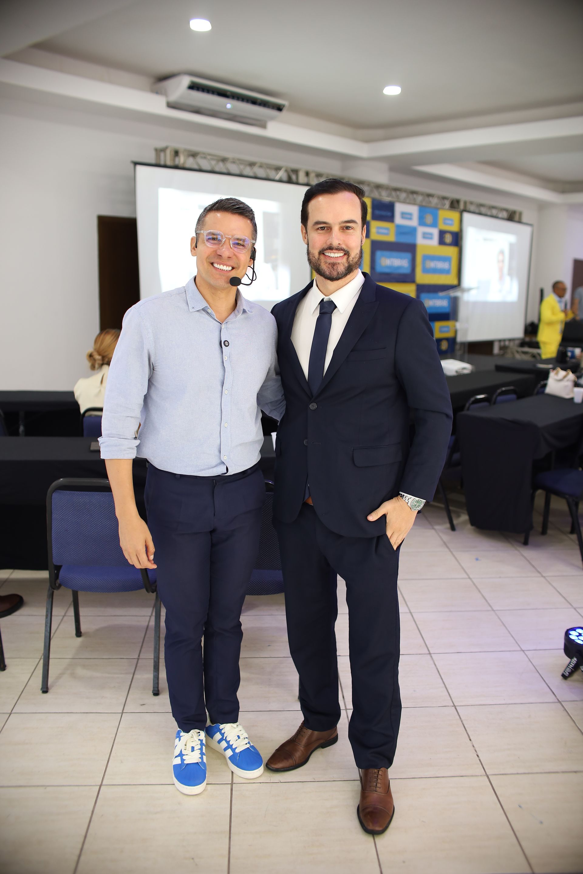 Dois homens sorrindo, posando juntos. Um de terno, o outro com roupa casual, em uma sala com uma tela de projeção.