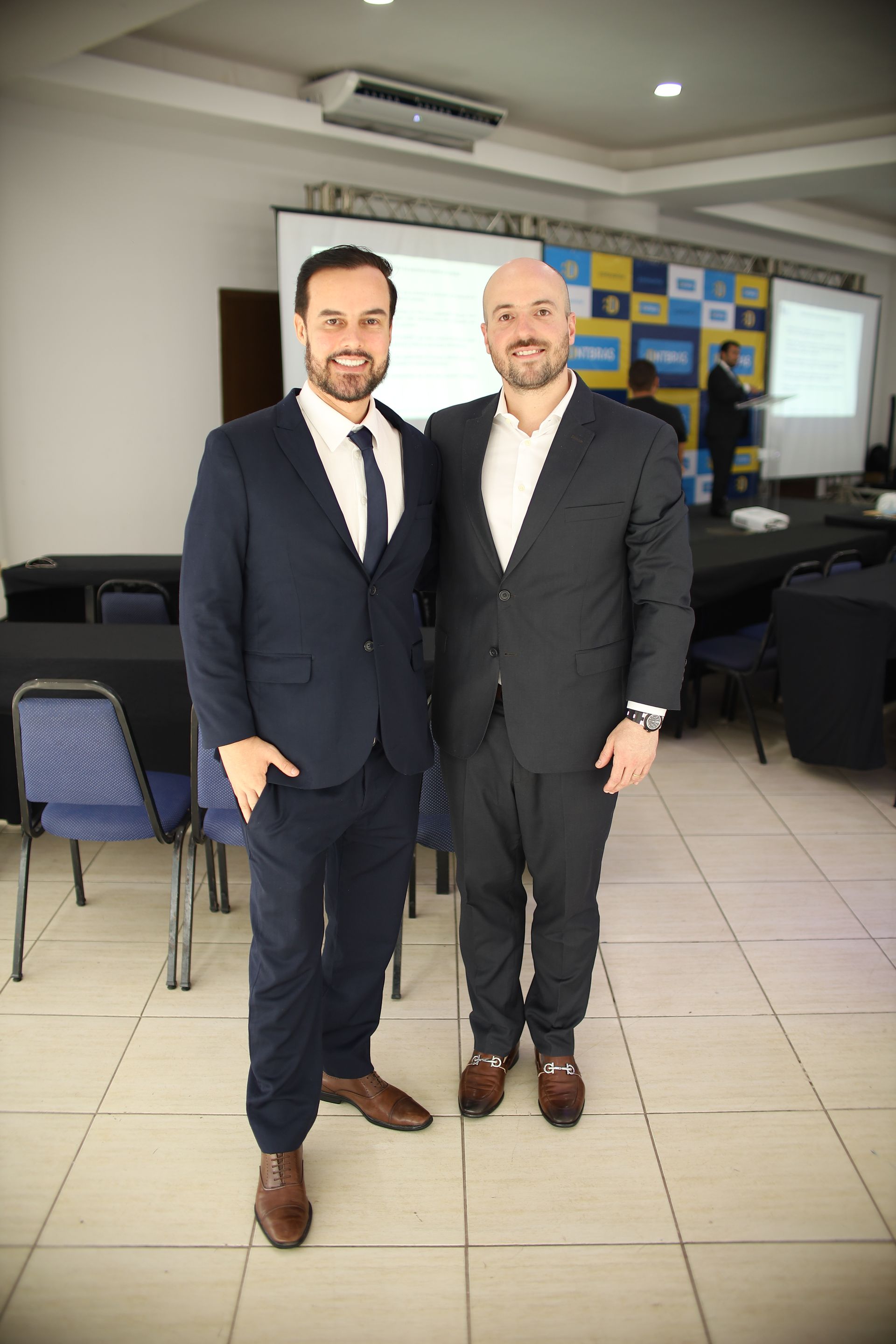 Dois homens de terno sorriem em uma sala de conferências com uma tela e mesas.