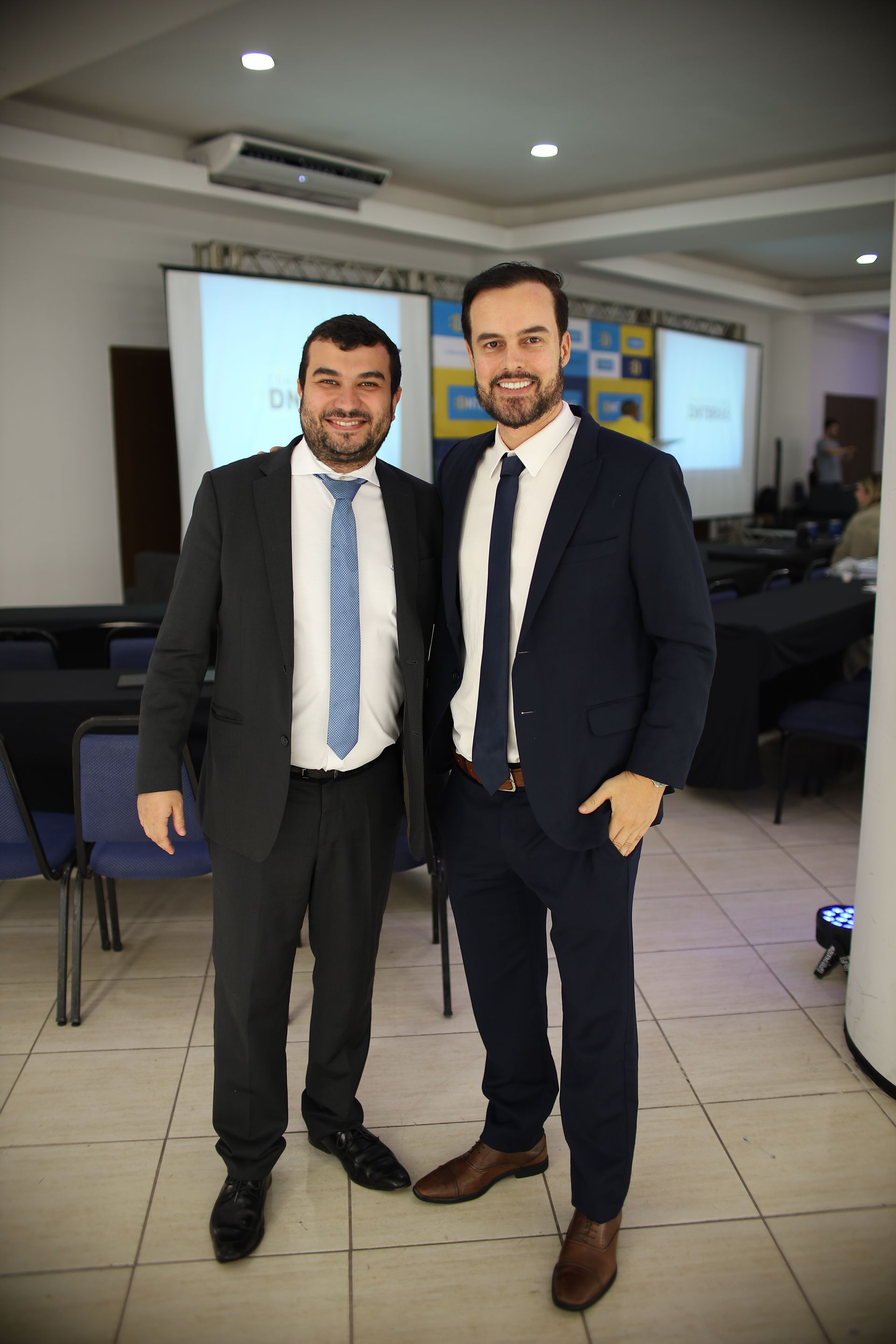 Dois homens de terno posam para fotos em um evento realizado em ambiente interno. A sala possui uma tela de projeção e mesas.
