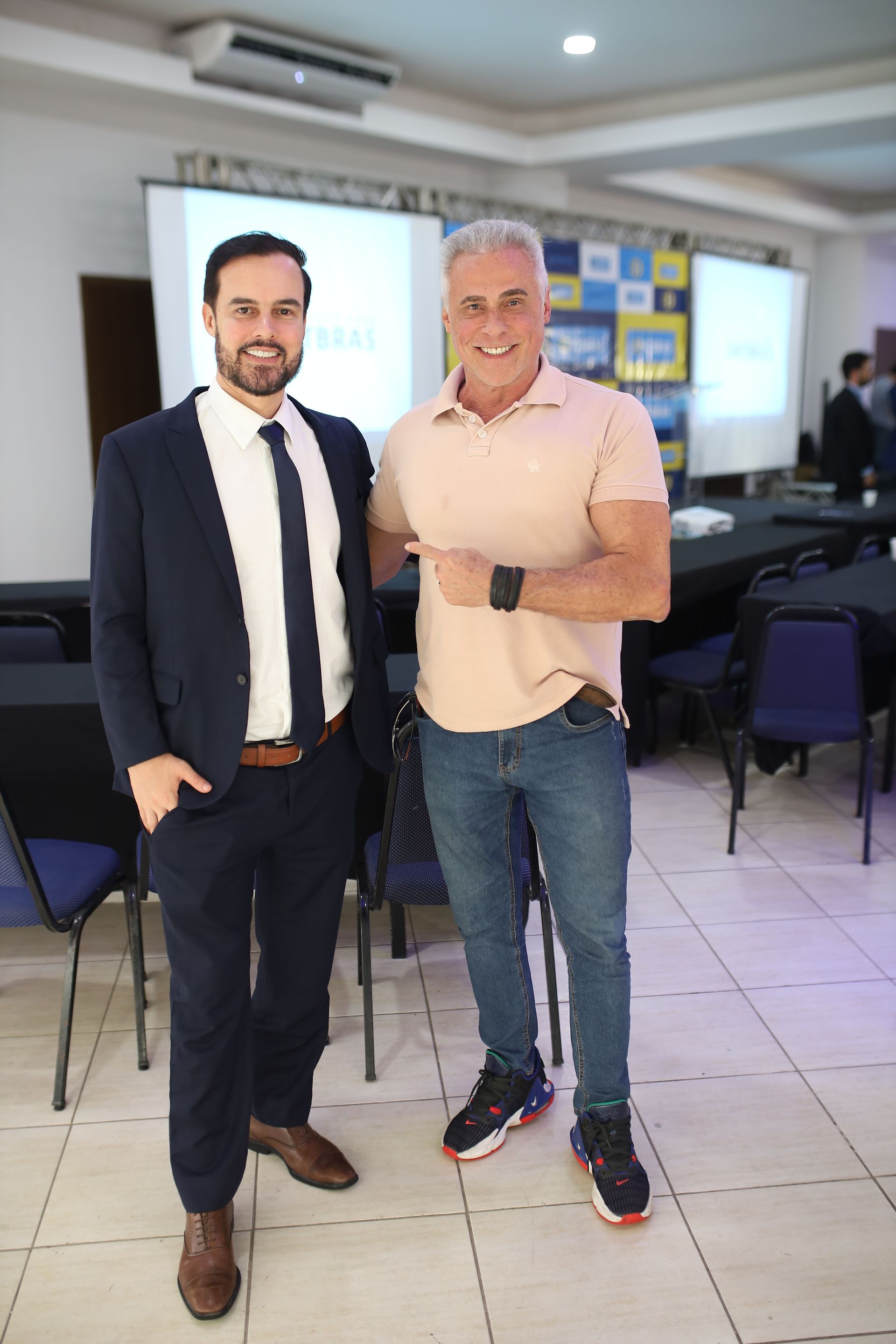 Dois homens posam para foto, um de terno e o outro de calça jeans e camiseta, em um evento com telão.
