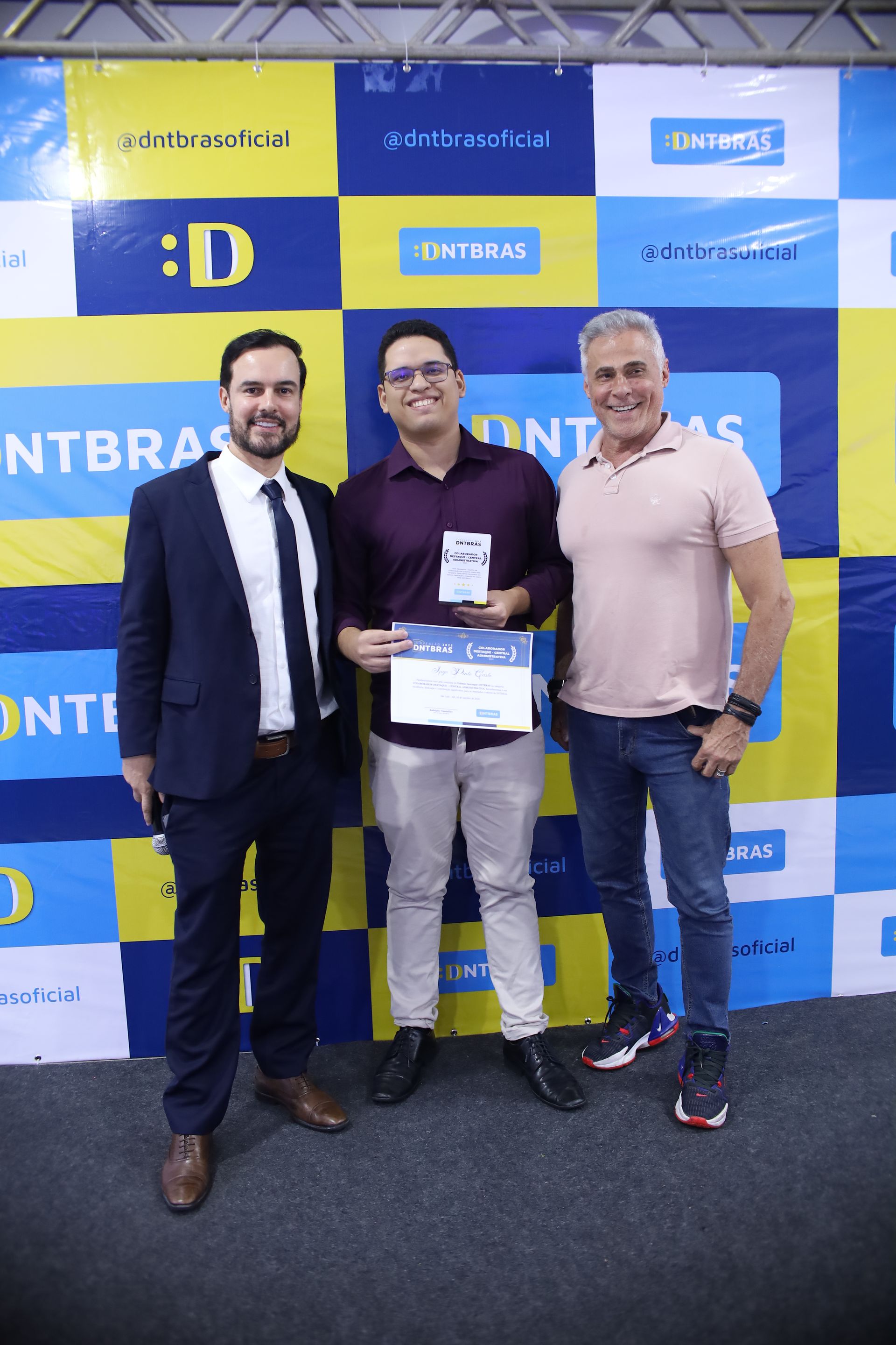 Três homens estão em pé diante de um fundo azul e amarelo com logotipos. O homem do meio segura um prêmio; eles sorriem.