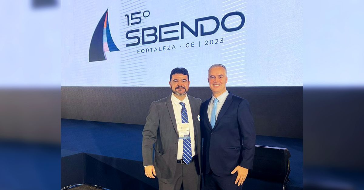 Dois homens de terno posam em frente a uma faixa da conferência SBENDO.