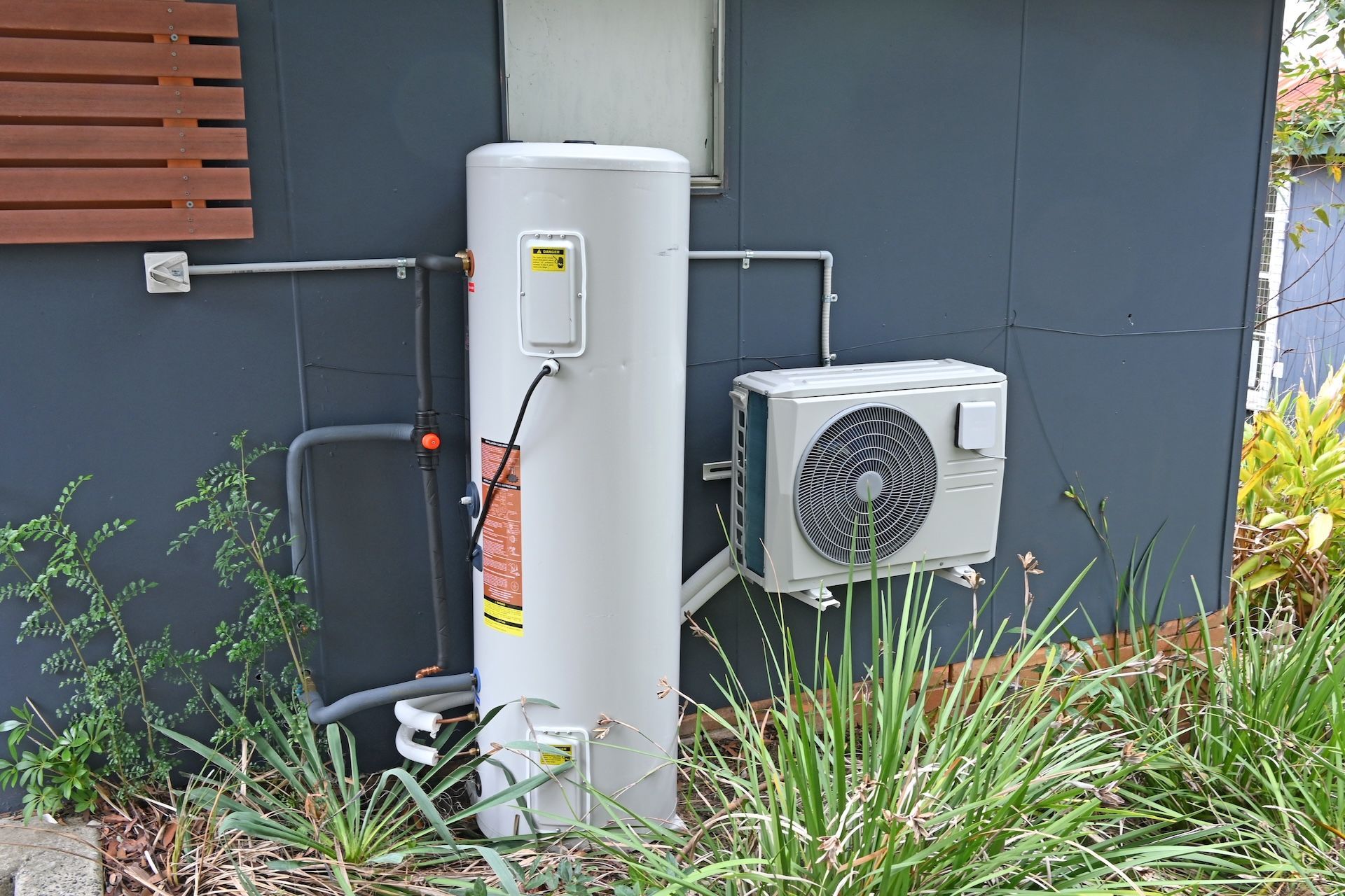 A white cylindrical heat pump water heater sits next to an outdoor air compressor unit against a dark grey exterior wall — Hot Water Specialist Forster In Forster, NSW