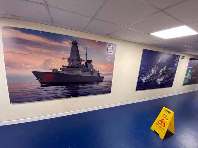 A caution sign is on the floor in a hallway with pictures of ships on the wall.