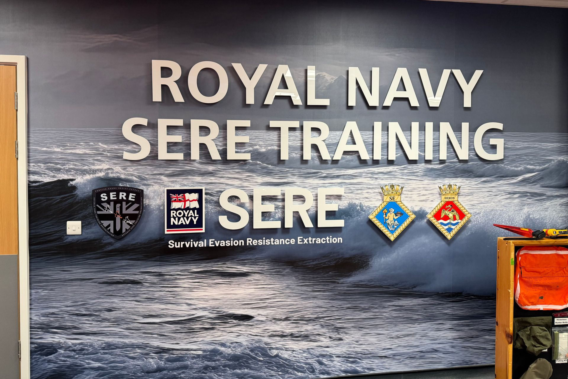 Royal Navy SERE Training sign with wave graphic and emblems.