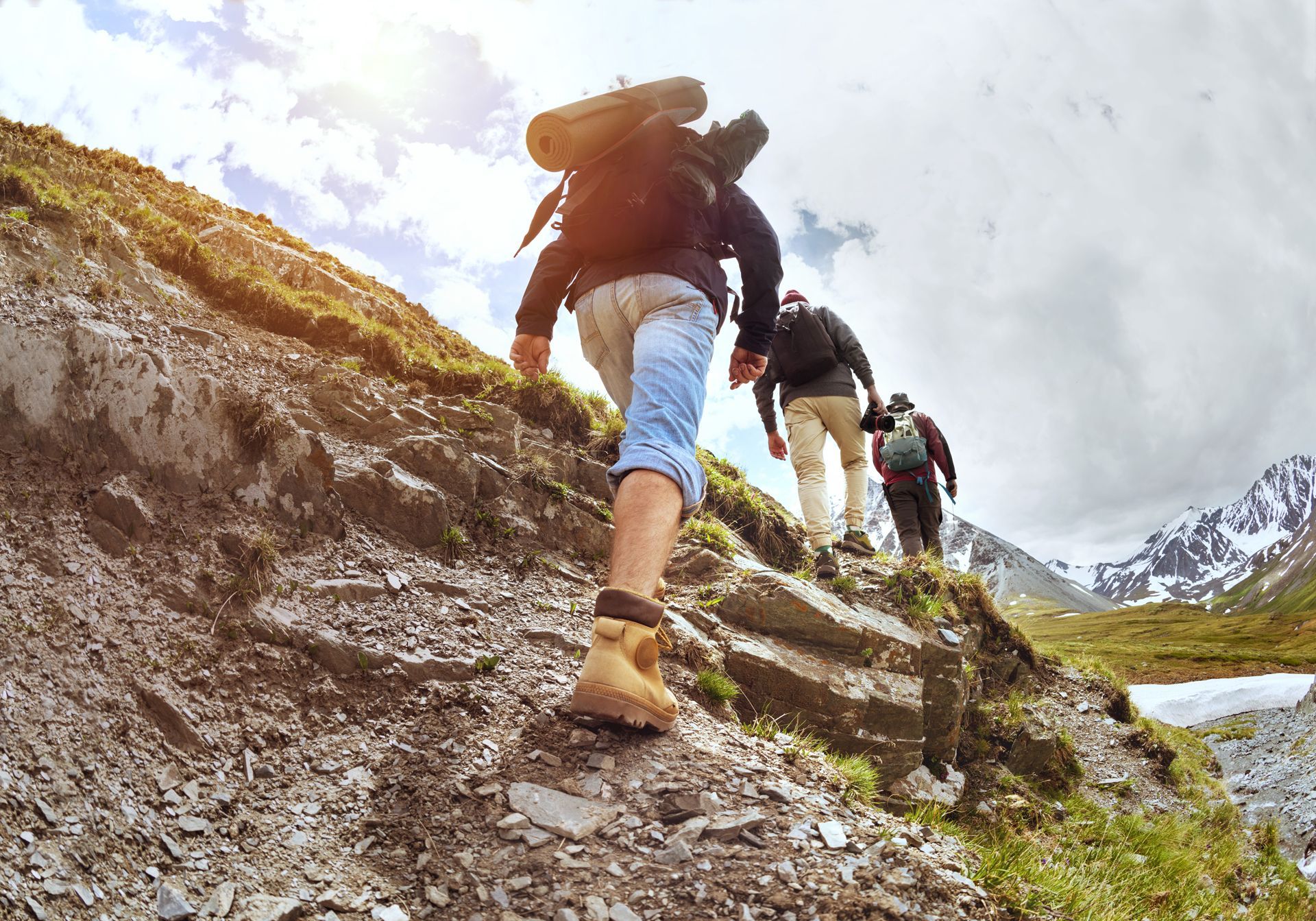 Three hikers ascending a rocky mountain trail, sunny sky, carrying backpacks.