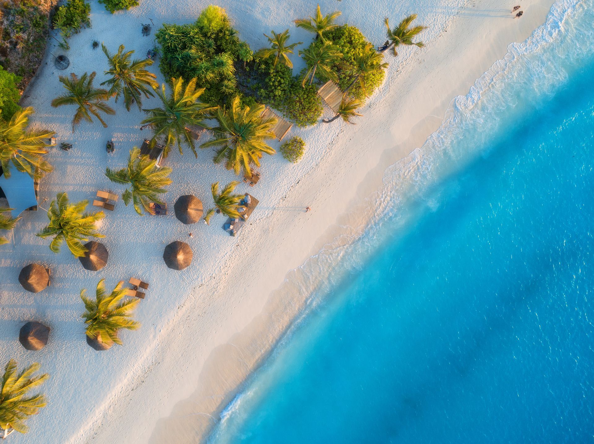 Aerial view of a tropical beach with turquoise water, white sand, palm trees, and thatched umbrellas