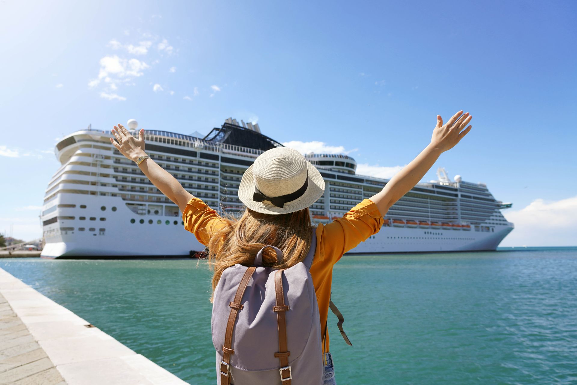 Woman with arms raised, facing a cruise ship, at a pier on a sunny day