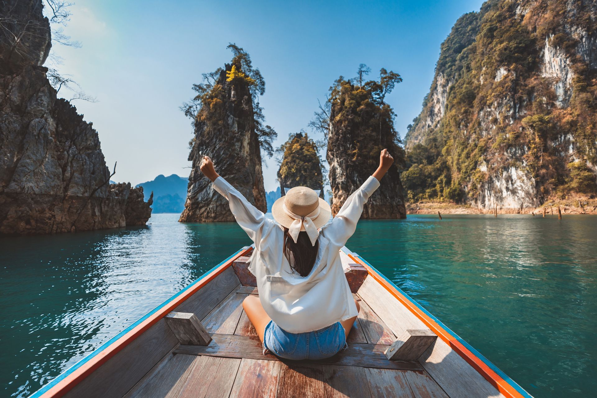 Woman in boat with arms raised, exploring emerald waters between rock formations under a clear blue sky