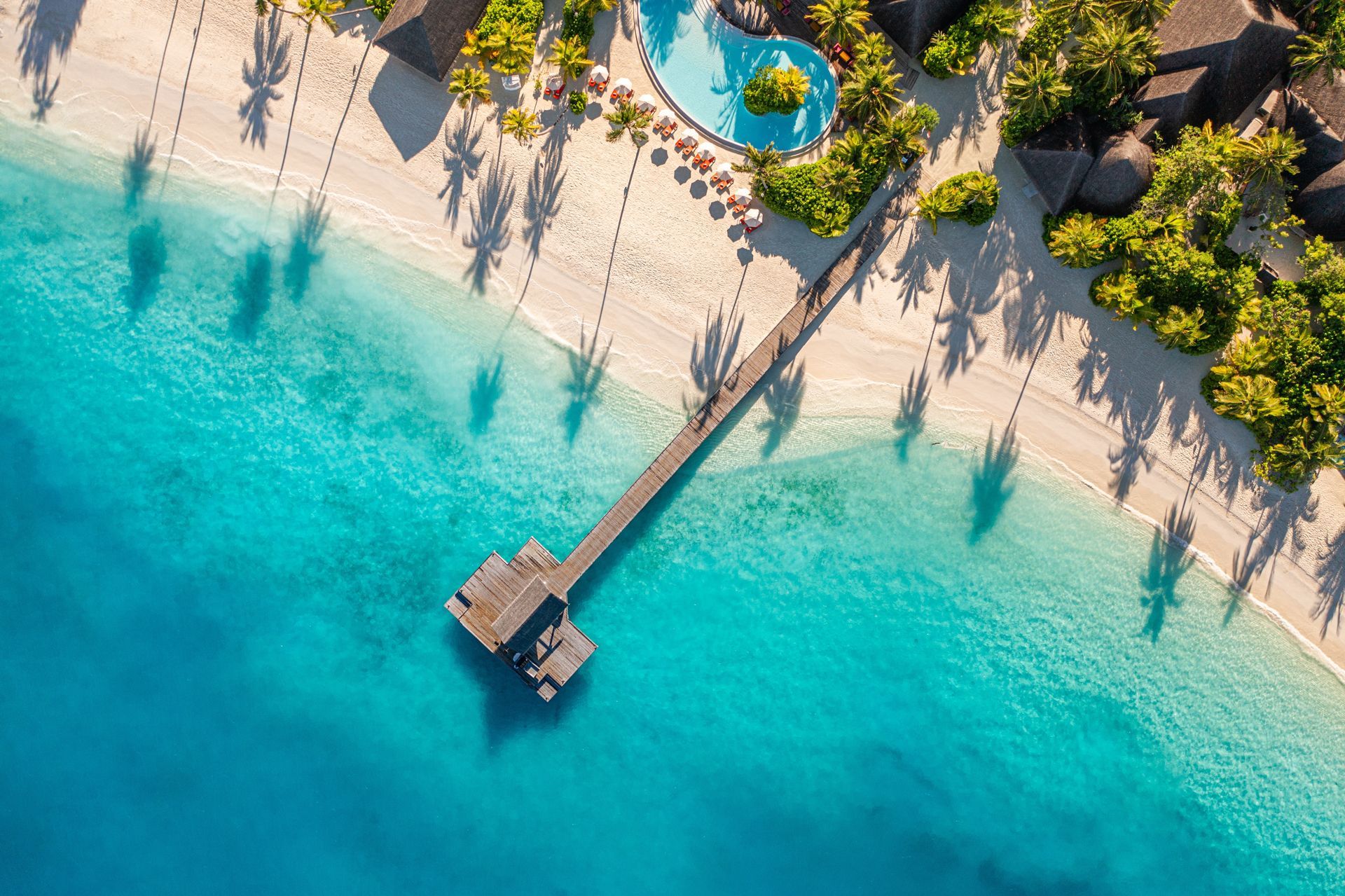 Aerial view: Pier extends into turquoise water, sandy beach, palm tree shadows, resort buildings
