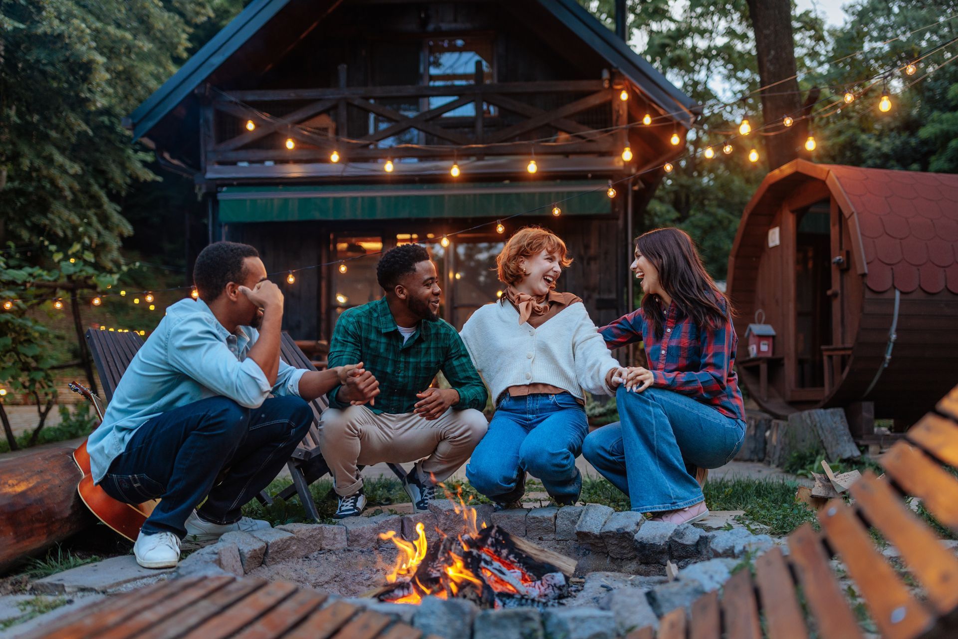 Four friends around a campfire in front of a cabin with string lights