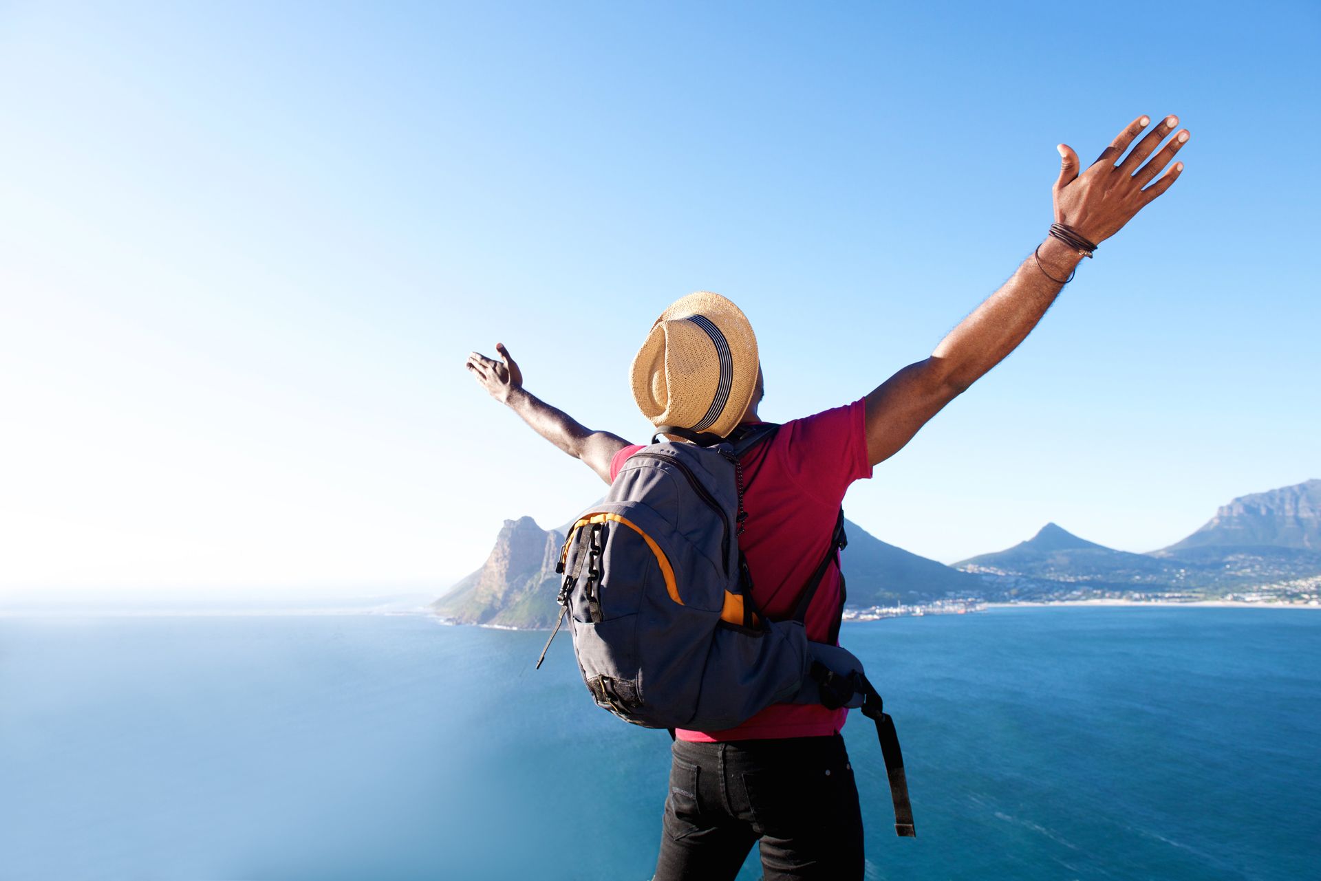 Man with backpack and hat raises arms, overlooking ocean and mountains on a sunny day
