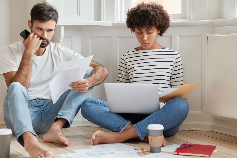 Couple sitting on floor, reviewing papers and using a laptop, looking concerned.