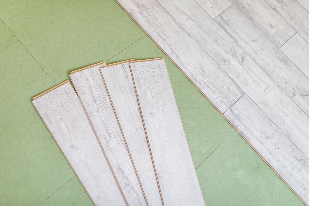 Laying laminate flooring: light wood planks on green underlayment, partially installed.