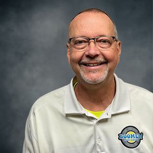 Smiling man in glasses and white pullover against a gray backdrop, wearing a logo on his chest.