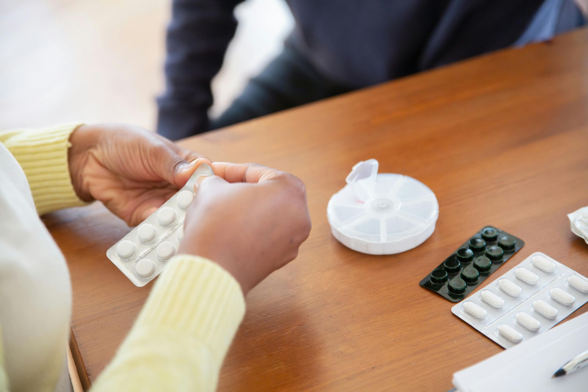 A woman is sitting at a table holding a blister pack of pills.