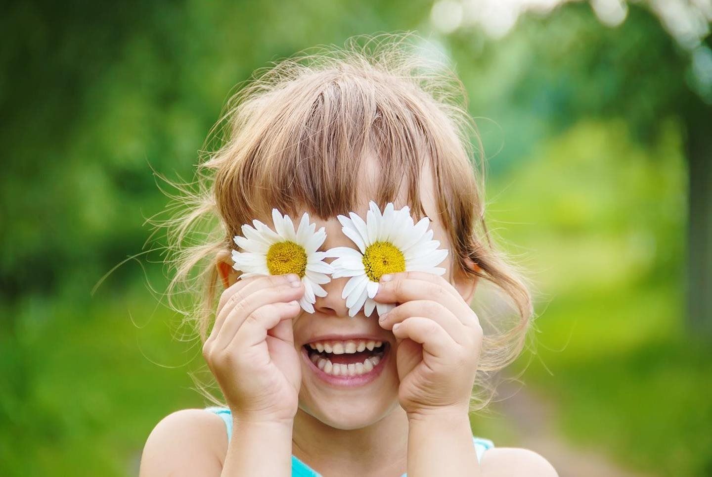 Smiling child holding daisies over their eyes in a green outdoor setting