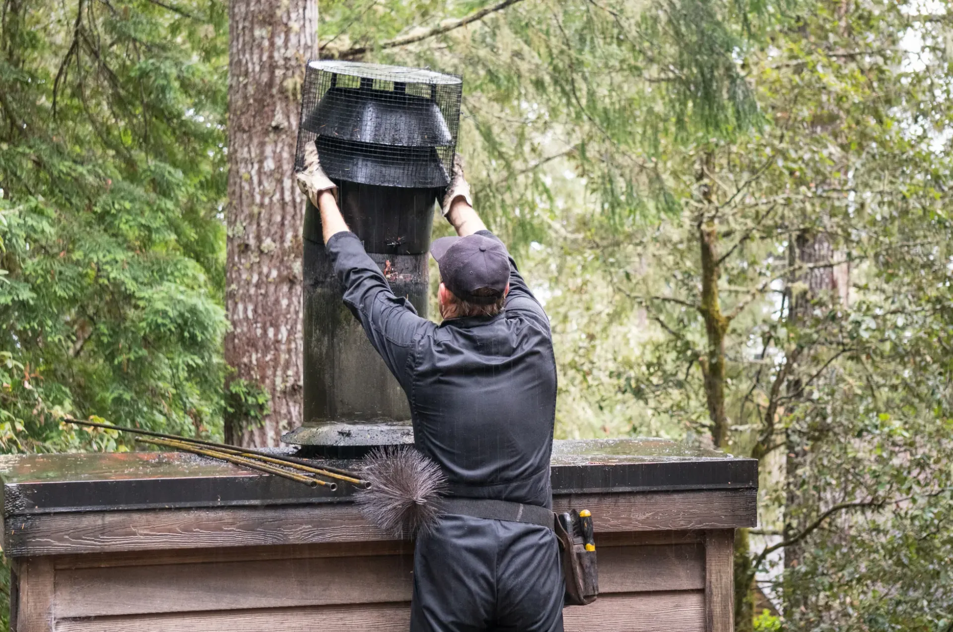 Man installs a chimney cap on a rooftop, set among trees.