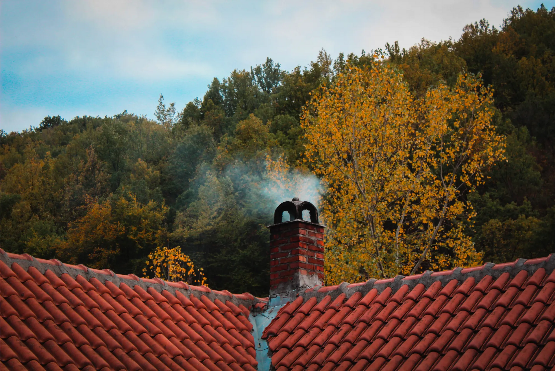 Chimney with smoke rising from red-tiled roof, backdrop of autumn trees against a blue sky.