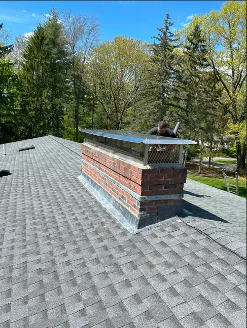 Chimney on a gray shingled roof with a metal cap. Trees and blue sky in the background.