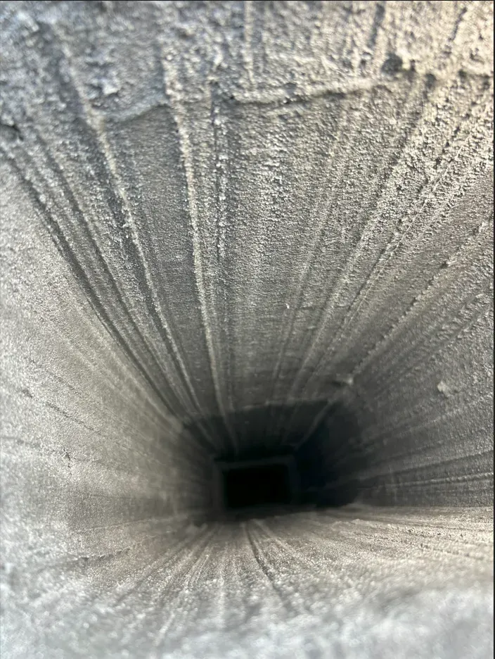 Interior view of a rectangular, dusty vent. Gray, fibrous material covers the walls, narrowing into the dark opening.