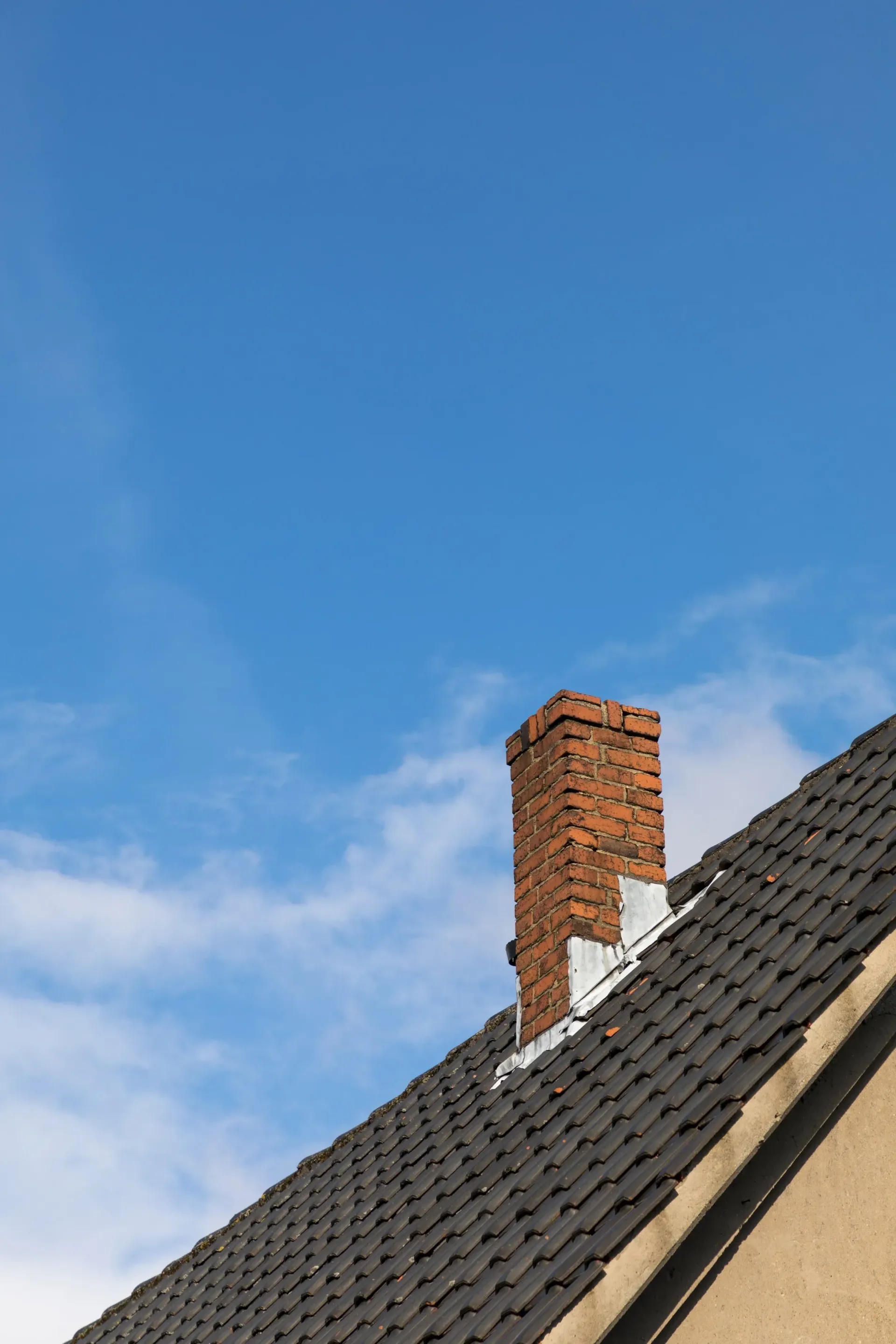Brick chimney on a dark-tiled roof against a blue sky with wispy clouds.