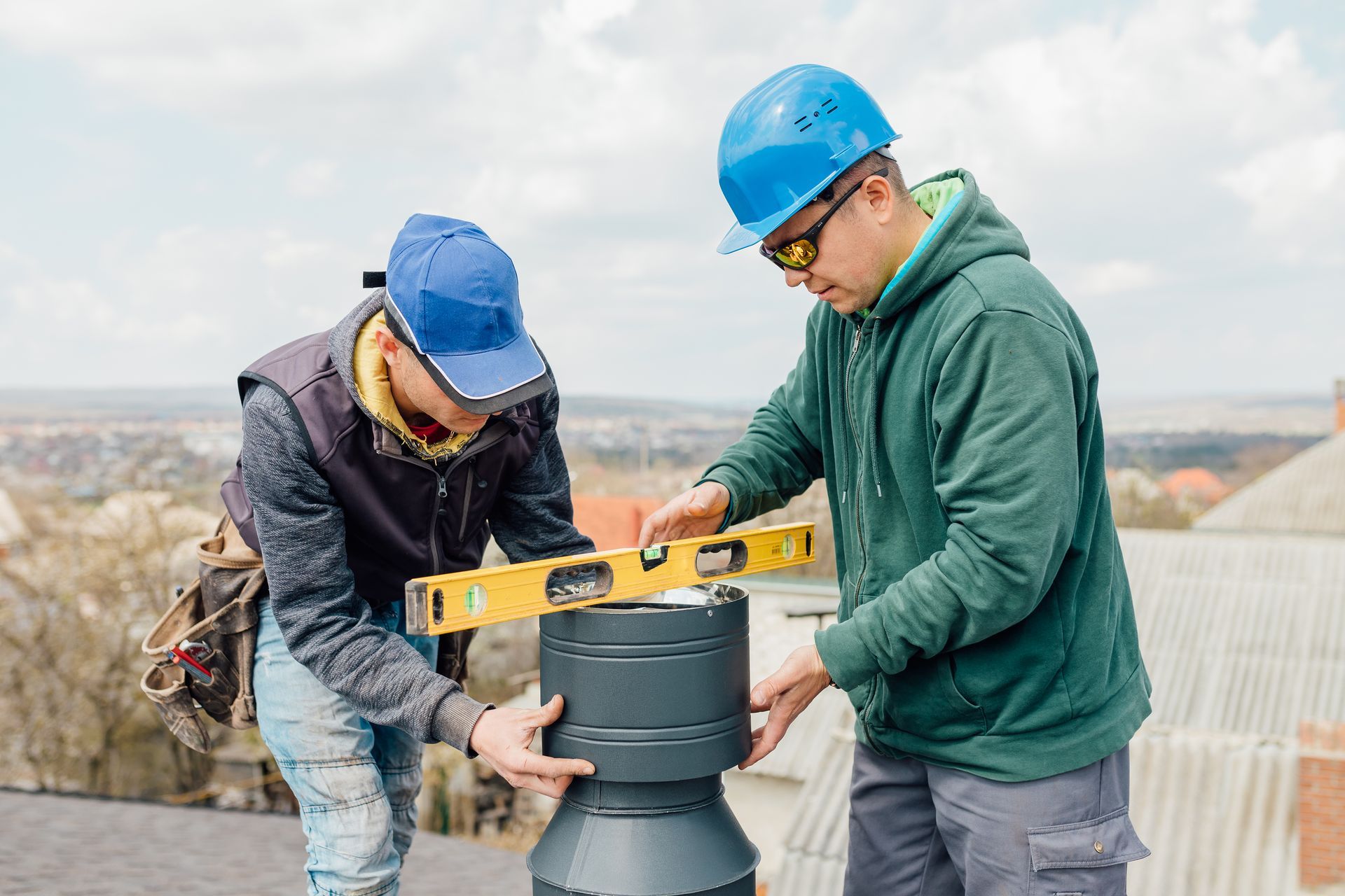 Two workers on a rooftop installing a chimney cap, using a level.