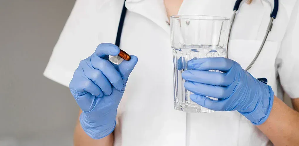 Person in medical attire holds a pill and a glass of water, preparing to administer medication.