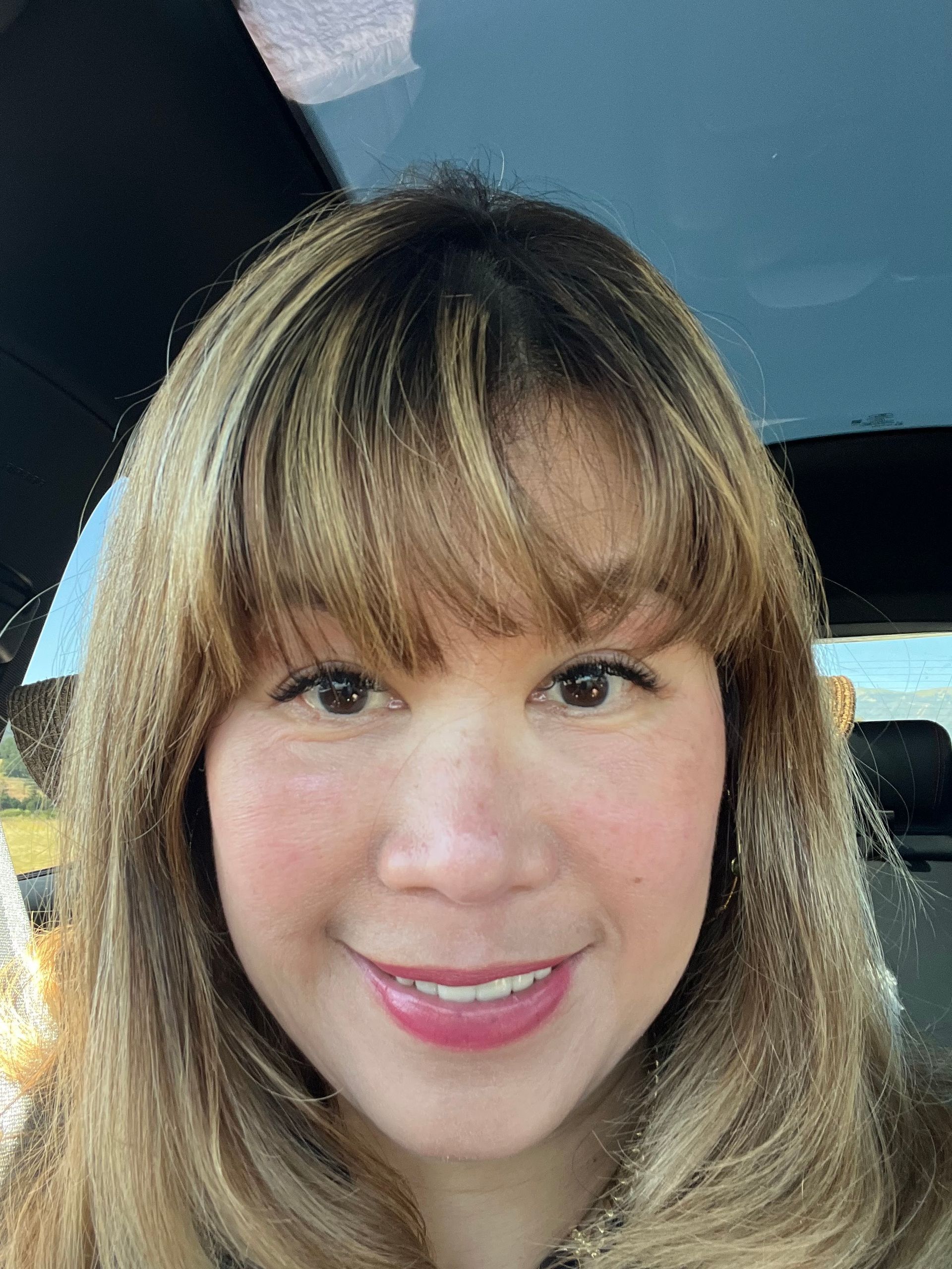 Woman with light brown hair and bangs smiles, rosy cheeks, possibly in a car.