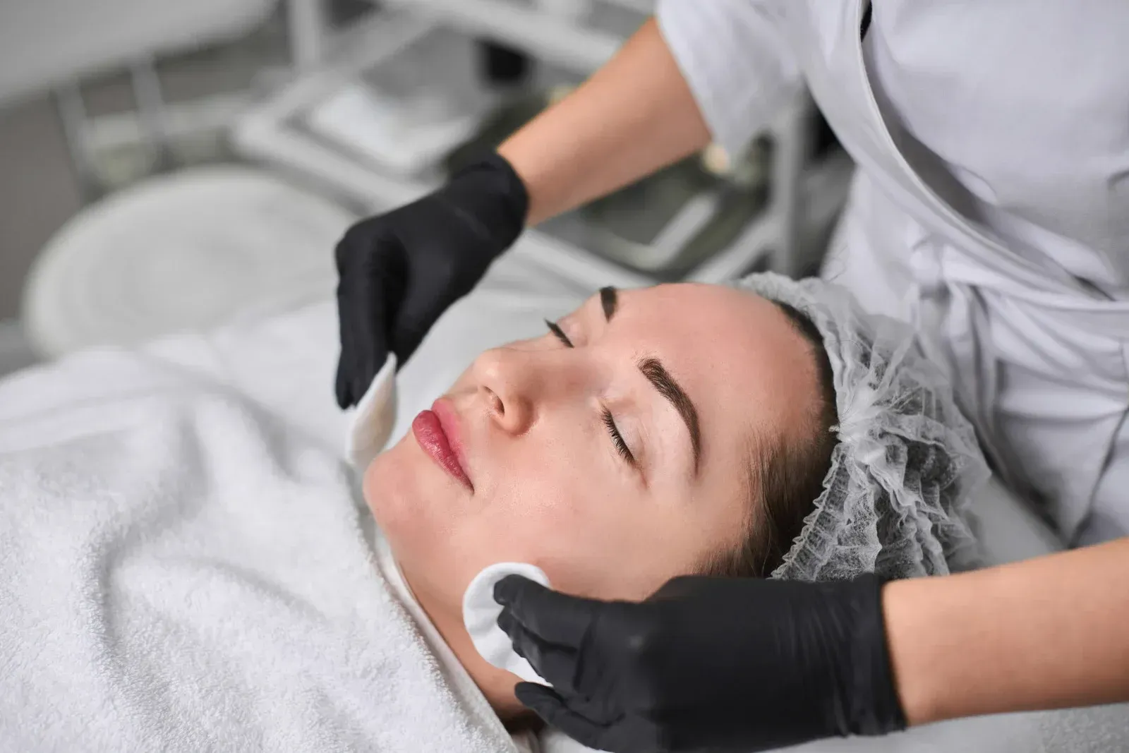 Woman receiving a facial treatment; esthetician applying product with cotton pads.