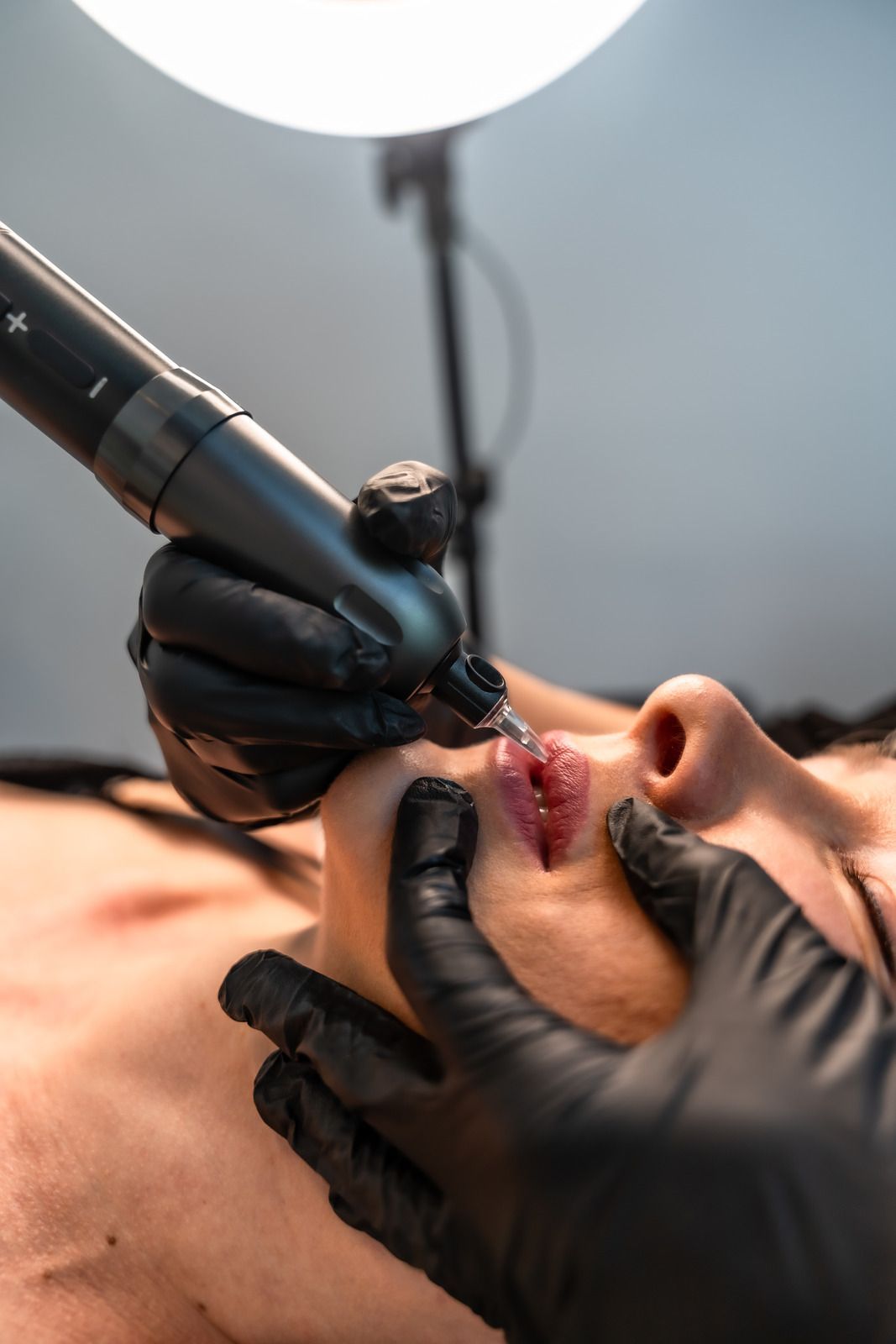 Person receiving lip tattoo; gloved hands, instrument applying pigment, bright light.