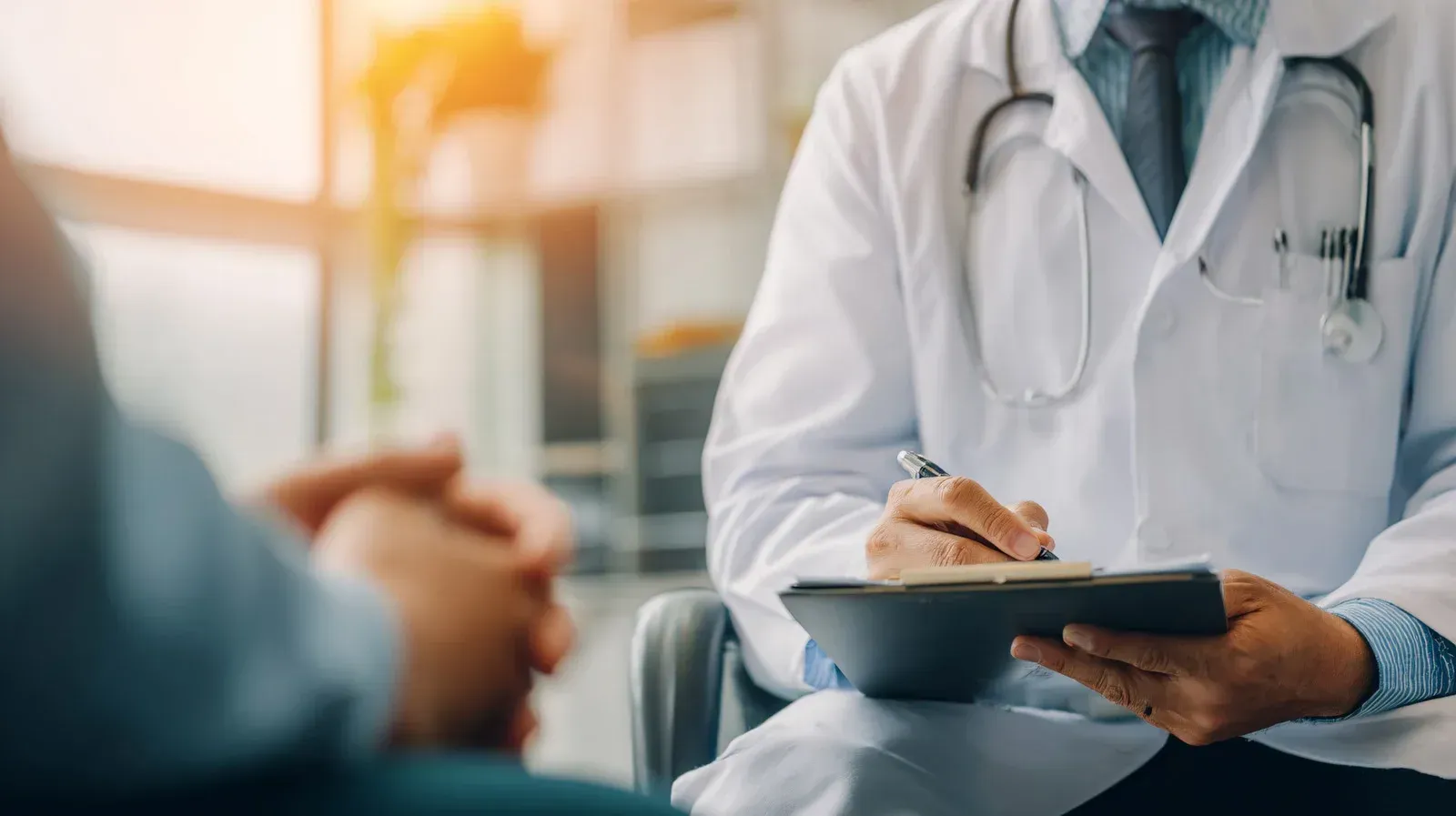 Doctor in white coat with stethoscope, taking notes on a clipboard while consulting with a patient.