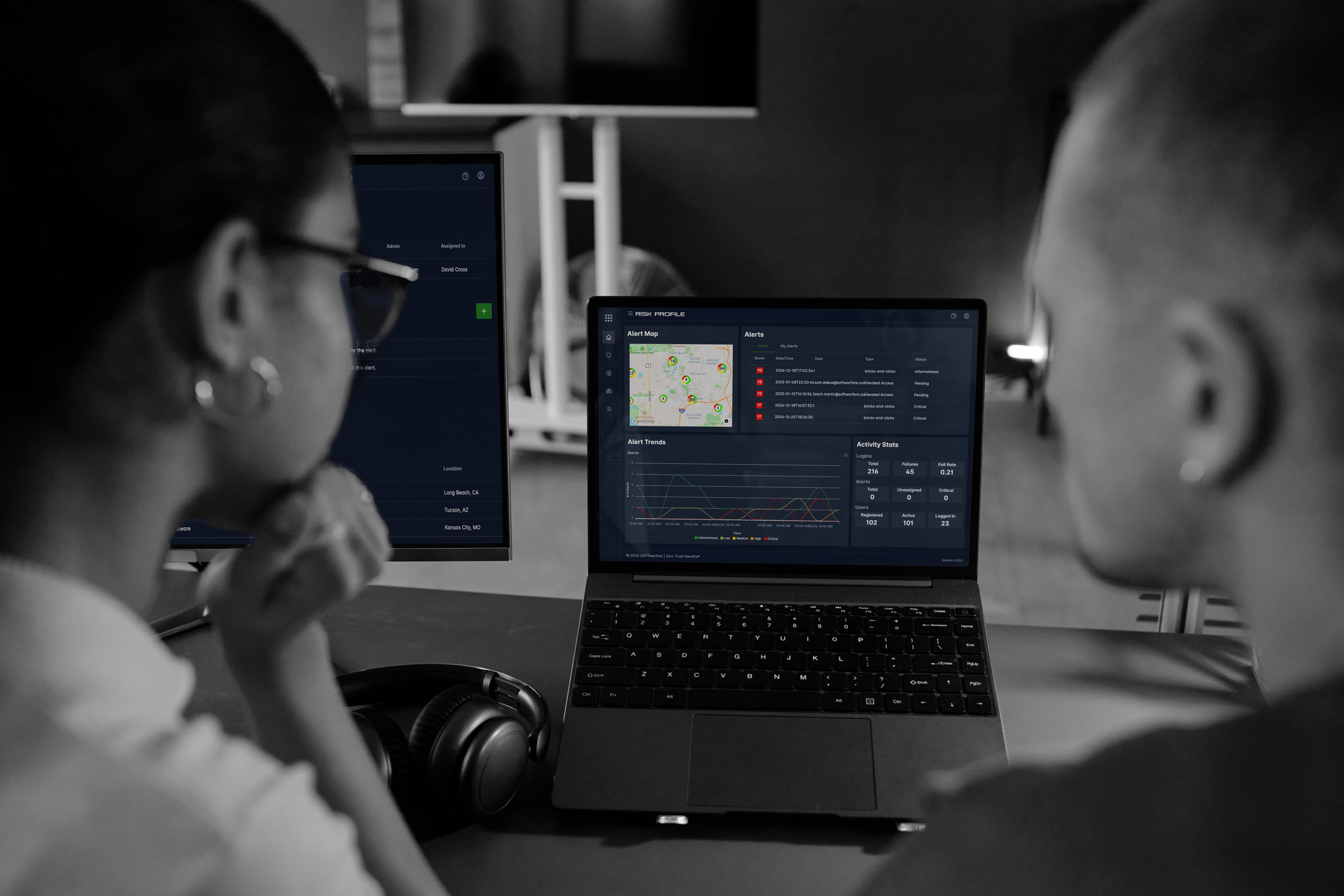 Two people looking at a laptop displaying data in a dark room. A vertical monitor sits behind the laptop.