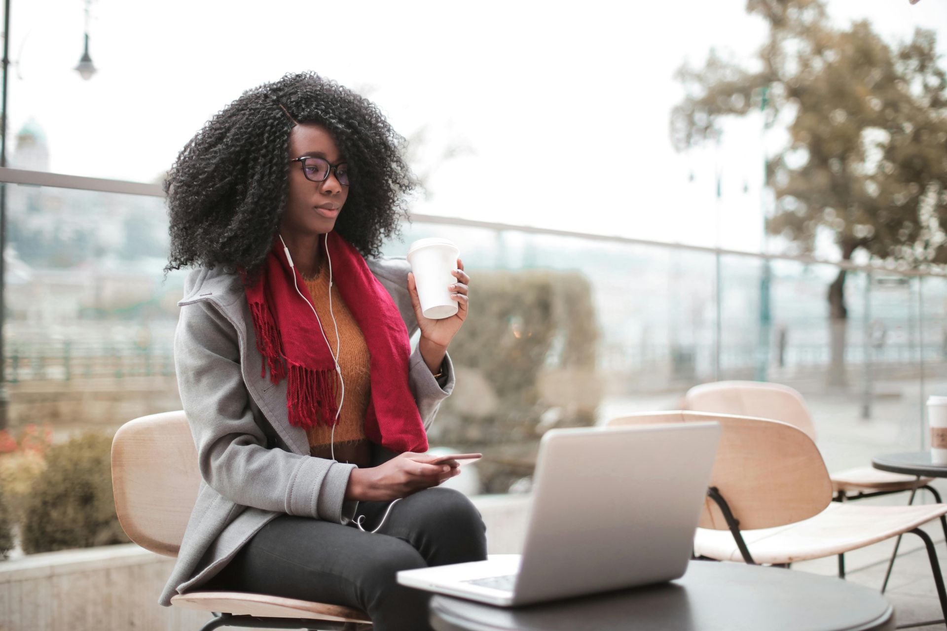 Woman with curly hair, glasses, and red scarf, working on a laptop, holding a coffee, outdoors.