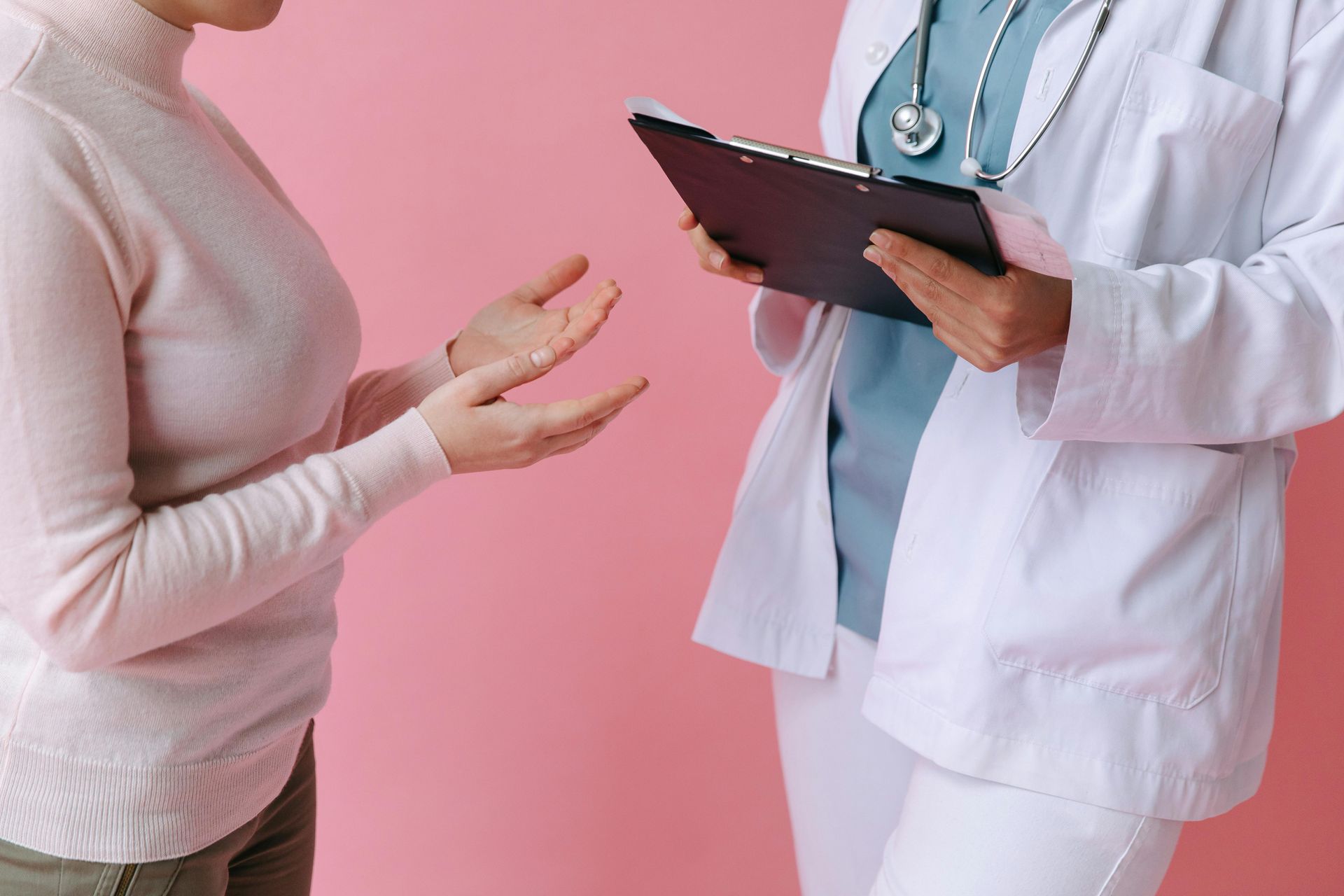 A patient gestures while conversing with a doctor in a white coat, pink background.