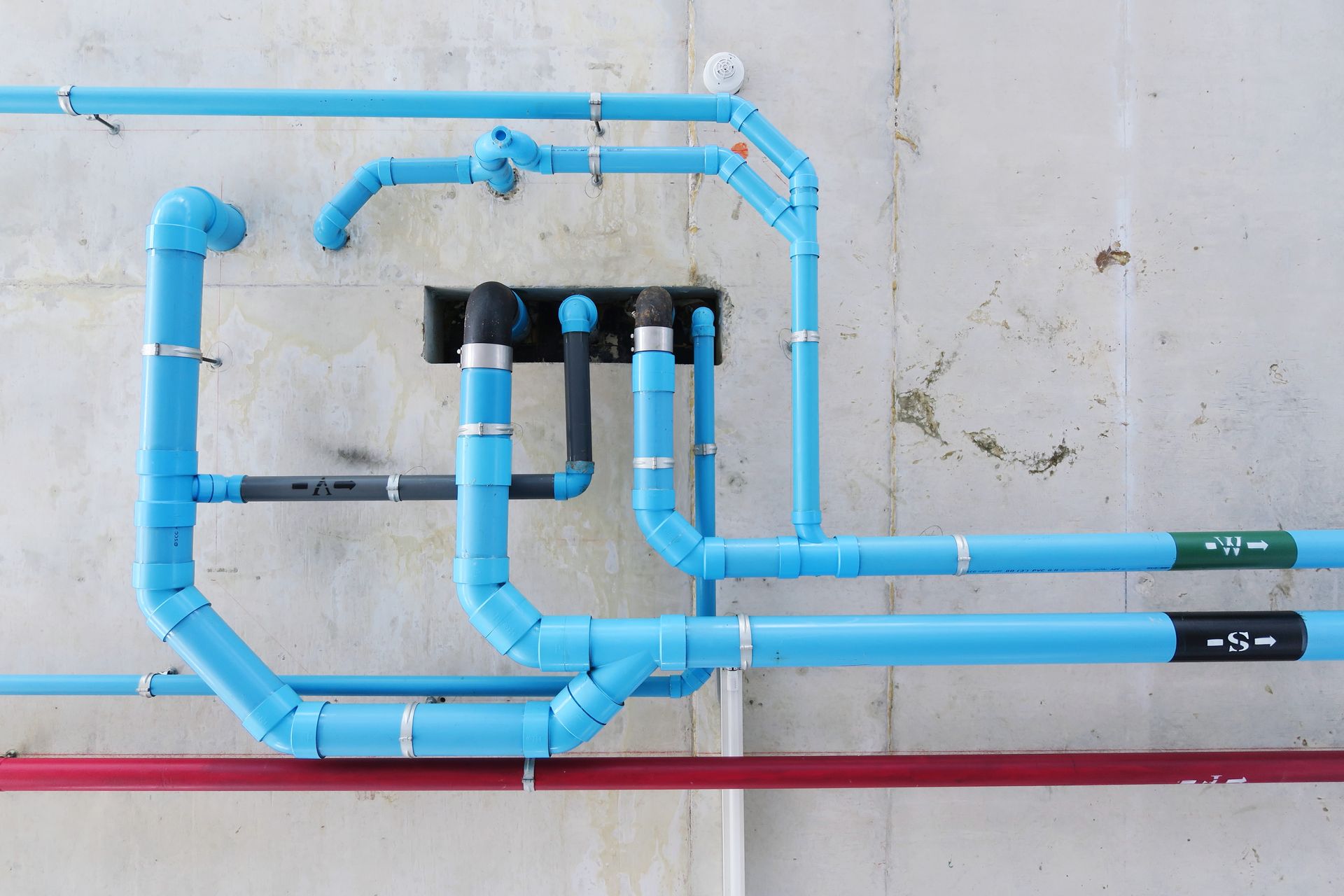 Blue PVC pipes and fittings on a concrete wall, with red pipe below.