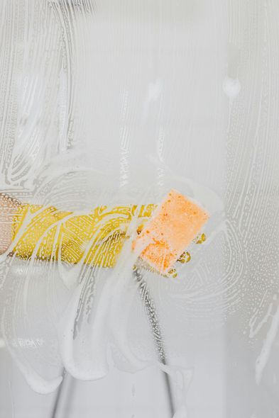 A hand with a sponge cleans a glass surface covered in soapy foam.