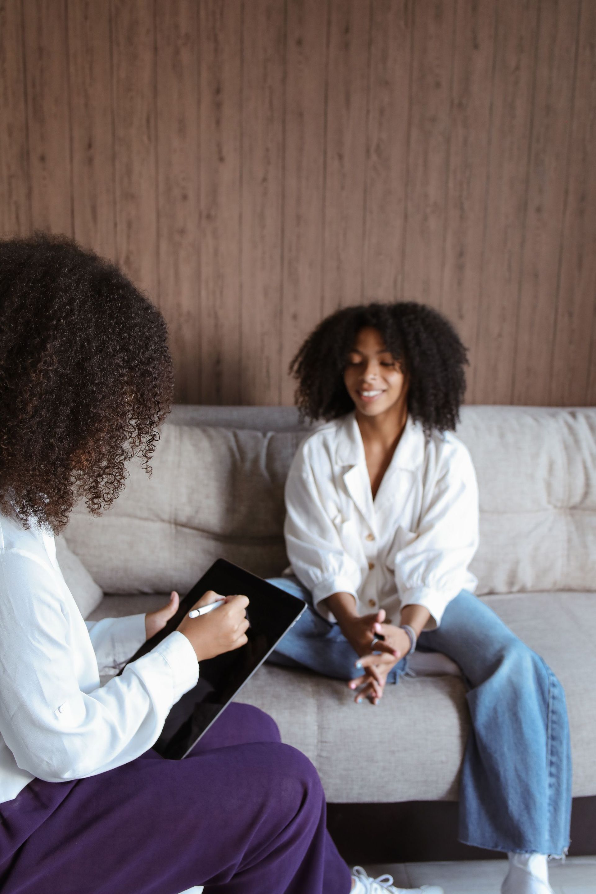Therapist taking notes while a seated client speaks on a beige couch in a counseling office