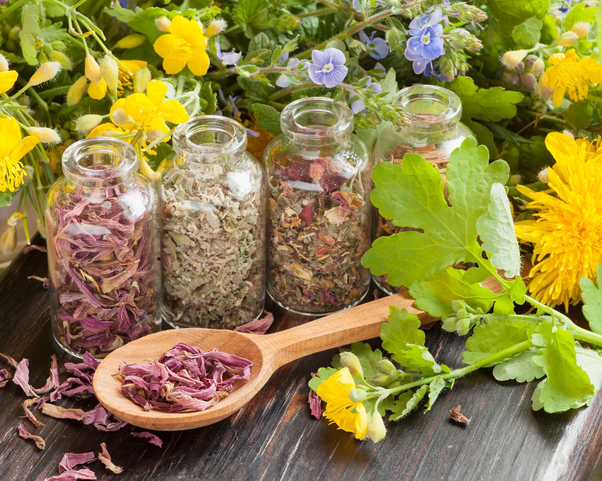 A wooden spoon filled with dried herbs next to bottles of herbs and flowers.
