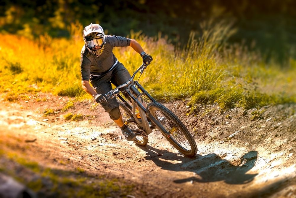 Mountain biker in gray gear leans into a turn on a dirt trail, sunlight glinting off the foliage.