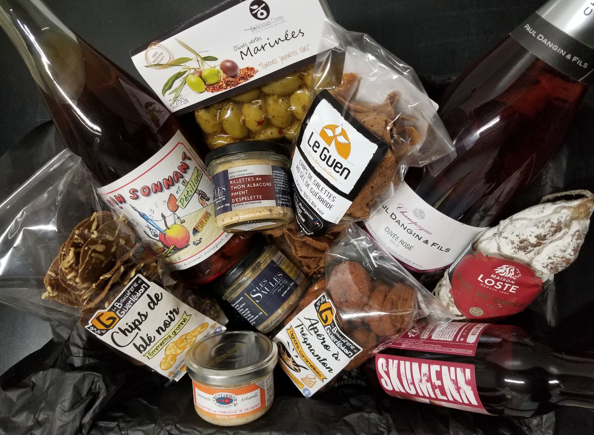 Panier-cadeau rempli de produits, notamment des olives, du pâté, de la charcuterie, des craquelins et des bouteilles de vin.