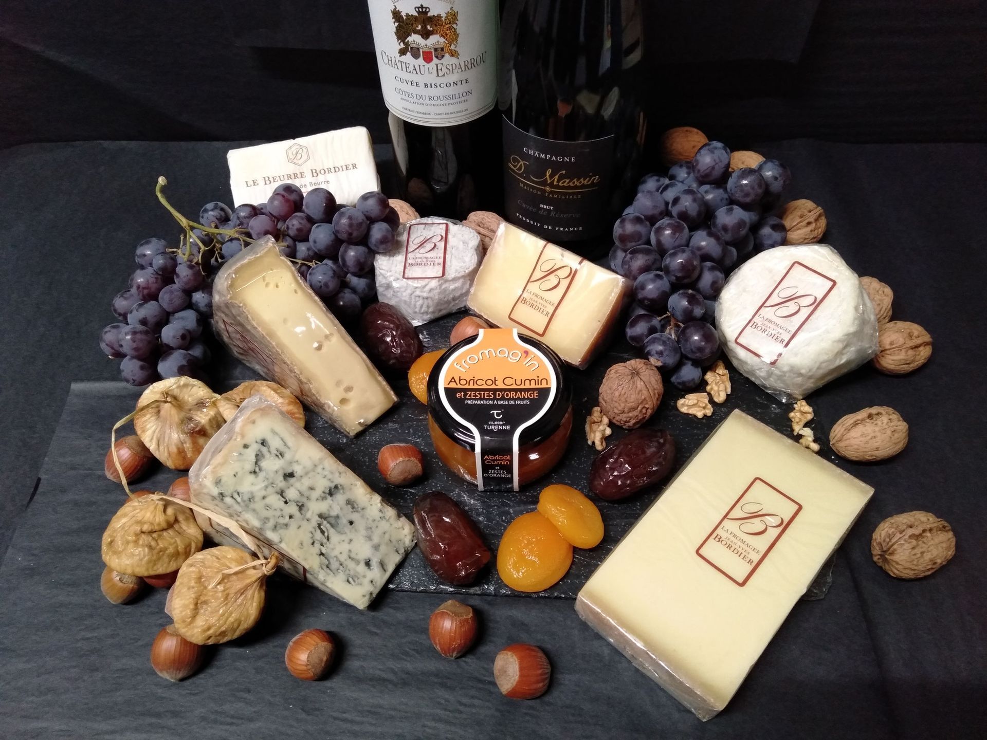 Plateau de fromages et de vins avec raisins, noix, dattes et abricots sur une surface en ardoise foncée.
