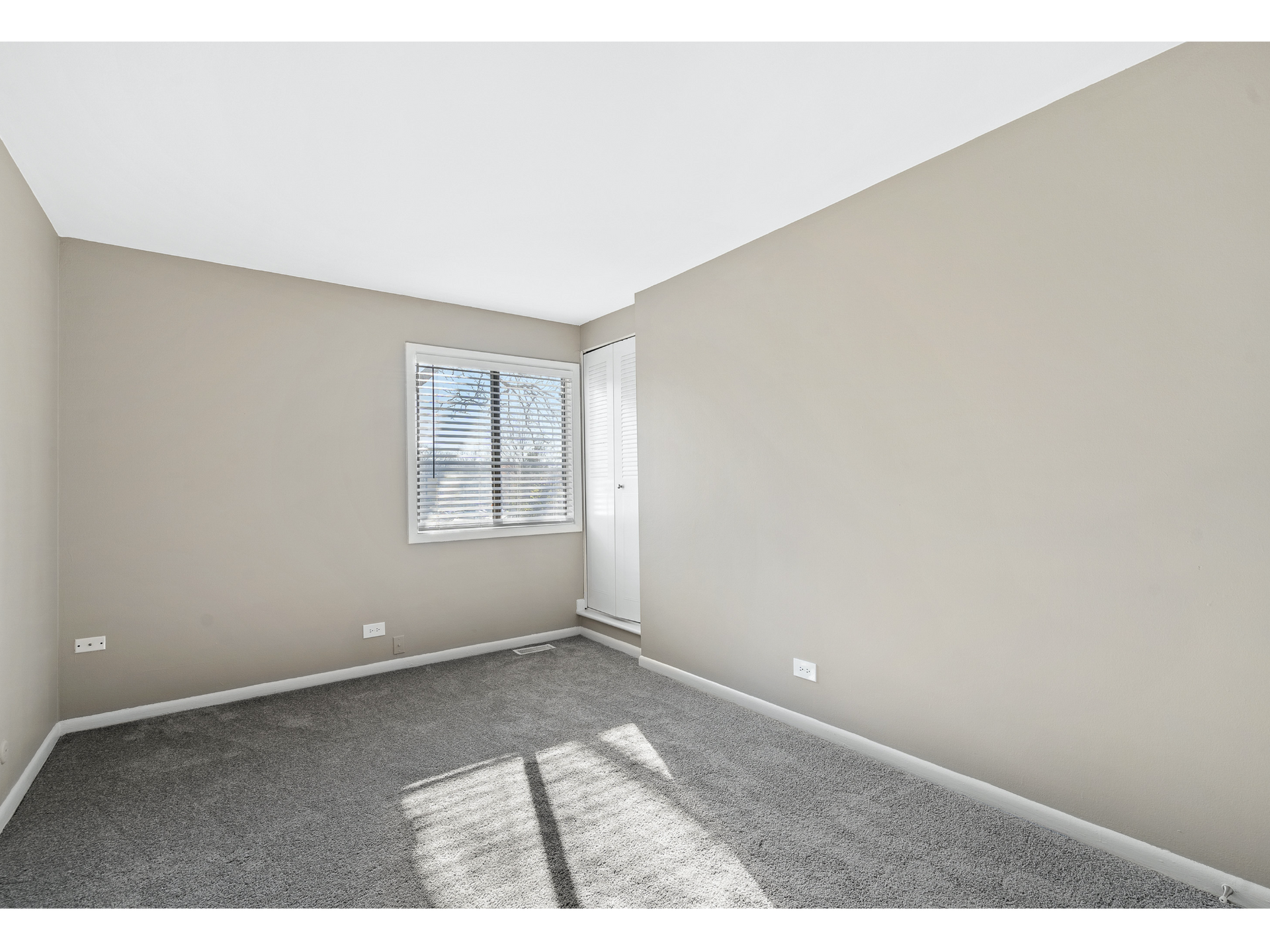 Empty beige bedroom with window, blinds, carpet, and a closet door.